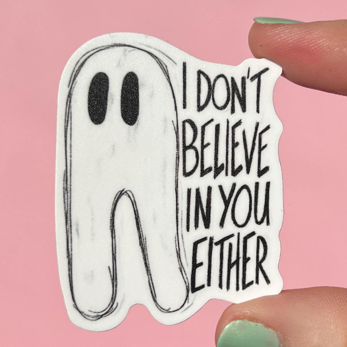 Sticker with ghost design and text 'I don't believe in you either' held by a hand against a pink background