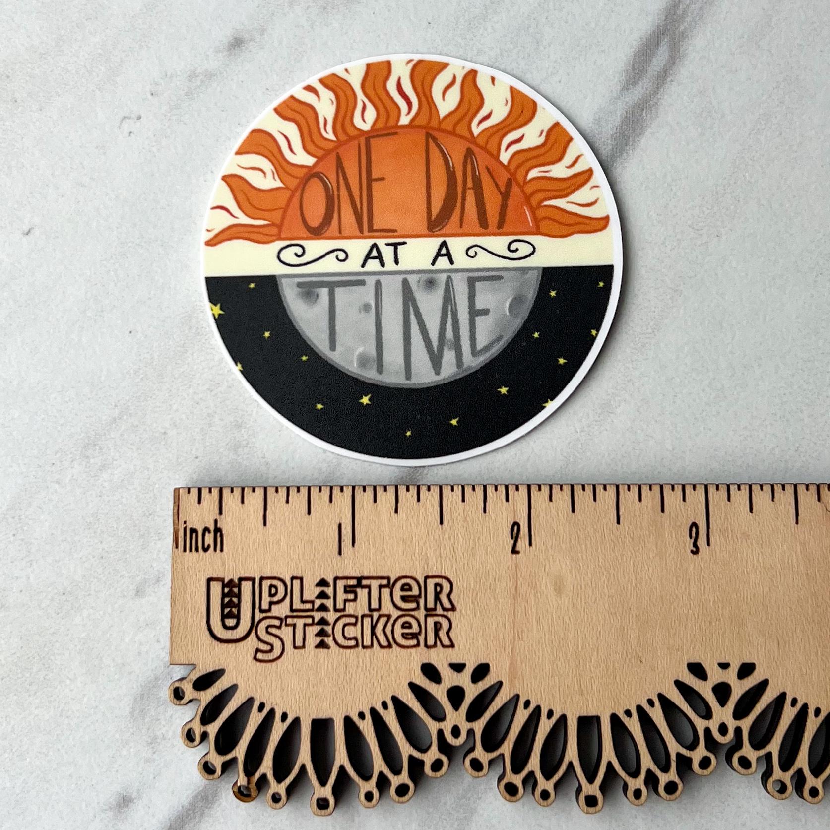 Circular sticker with 'One Day at a Time' design next to a ruler for size reference