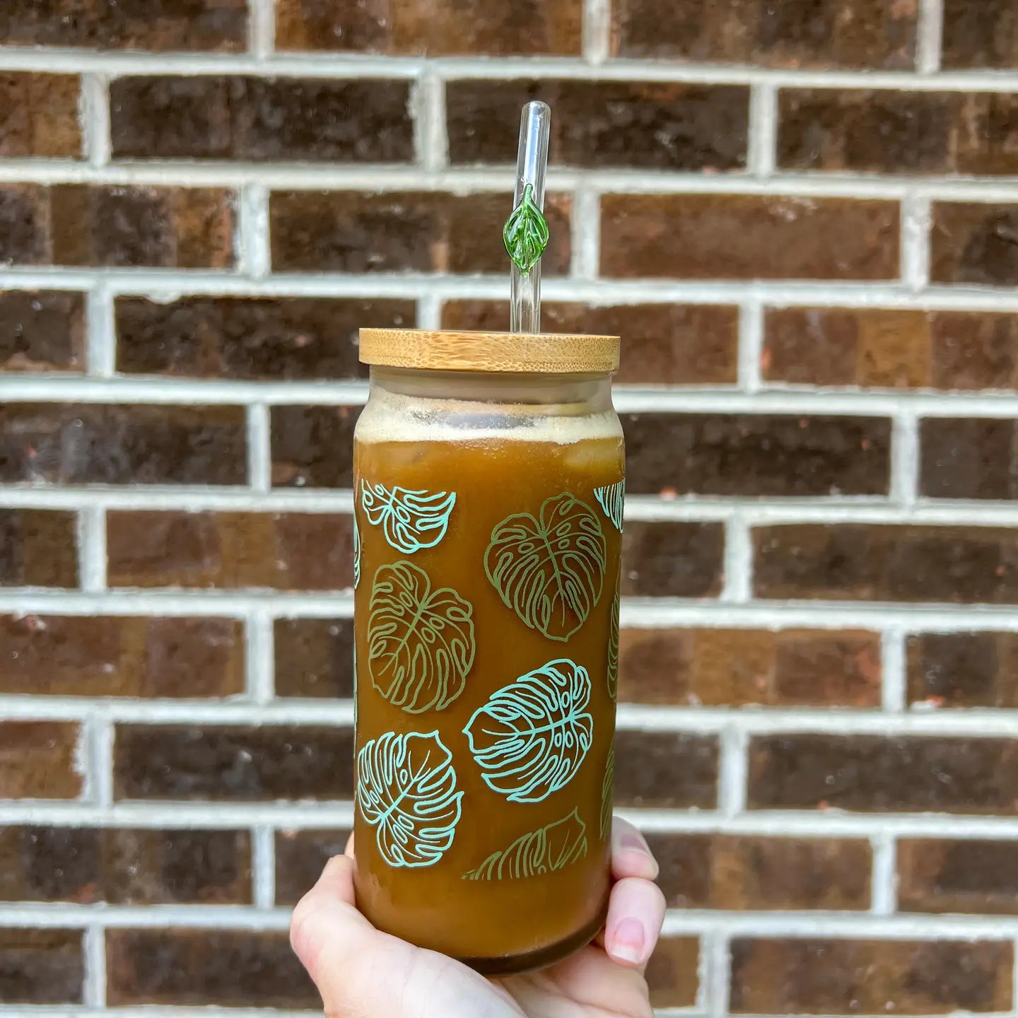 Hand holding a glass with a monstera cup pattern against a brick wall