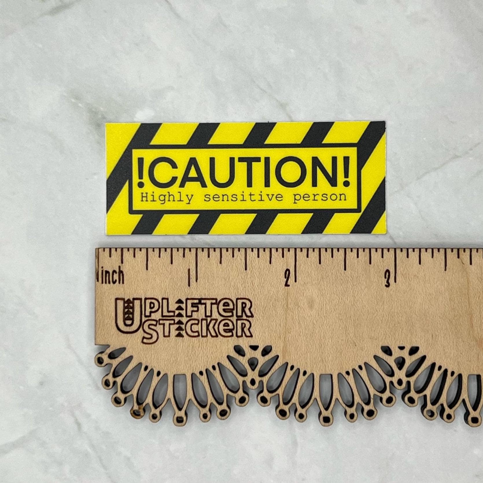 Caution sticker stating "caution highly sensitive person" with black and yellow stripes and text, placed on a ruler for size reference.
