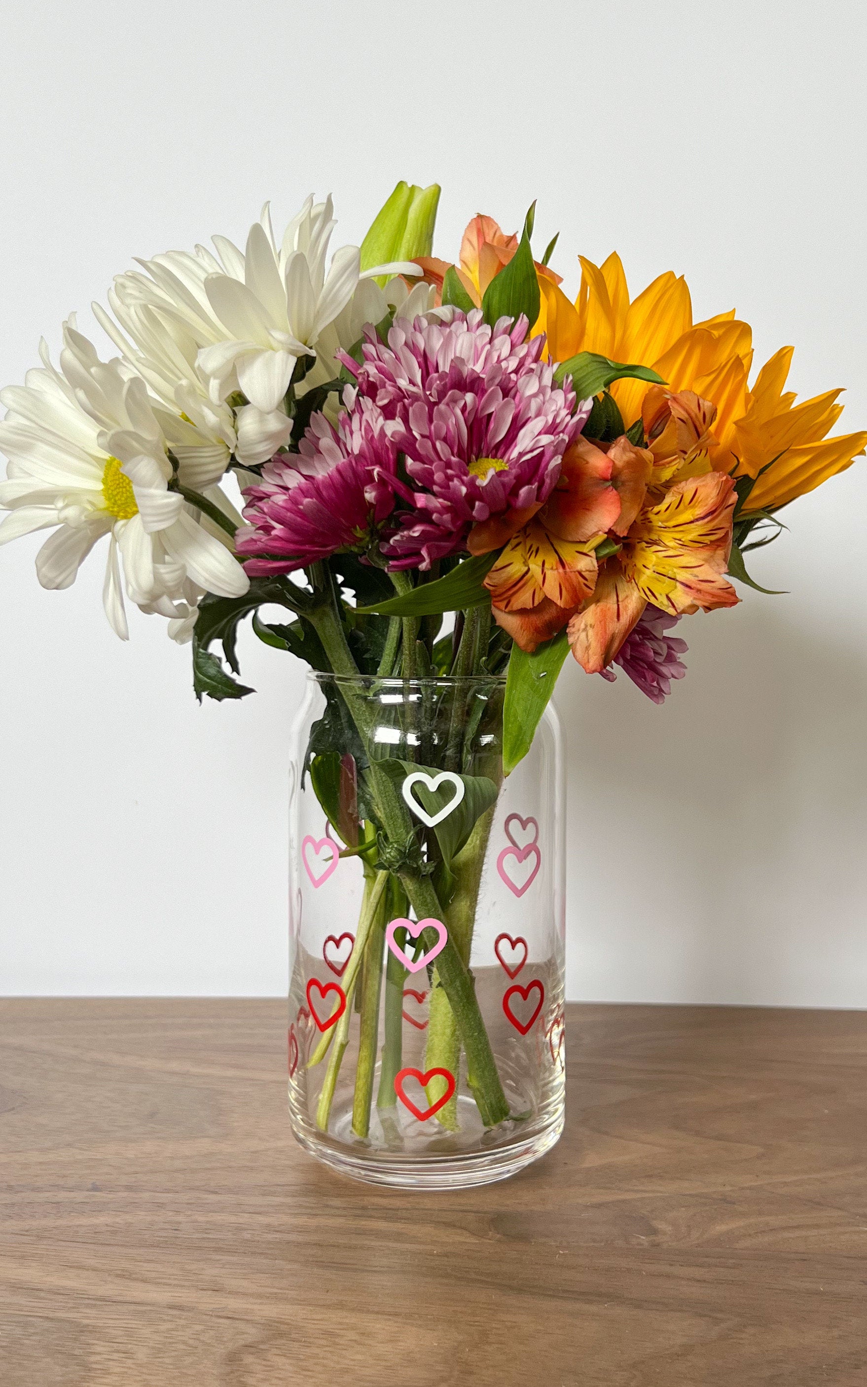 Clear vase with white, pink, and red heart designs filled with colorful flowers on a wooden surface.