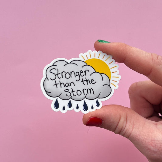 Sticker with 'Stronger than the storm' text held by a hand against a pink background