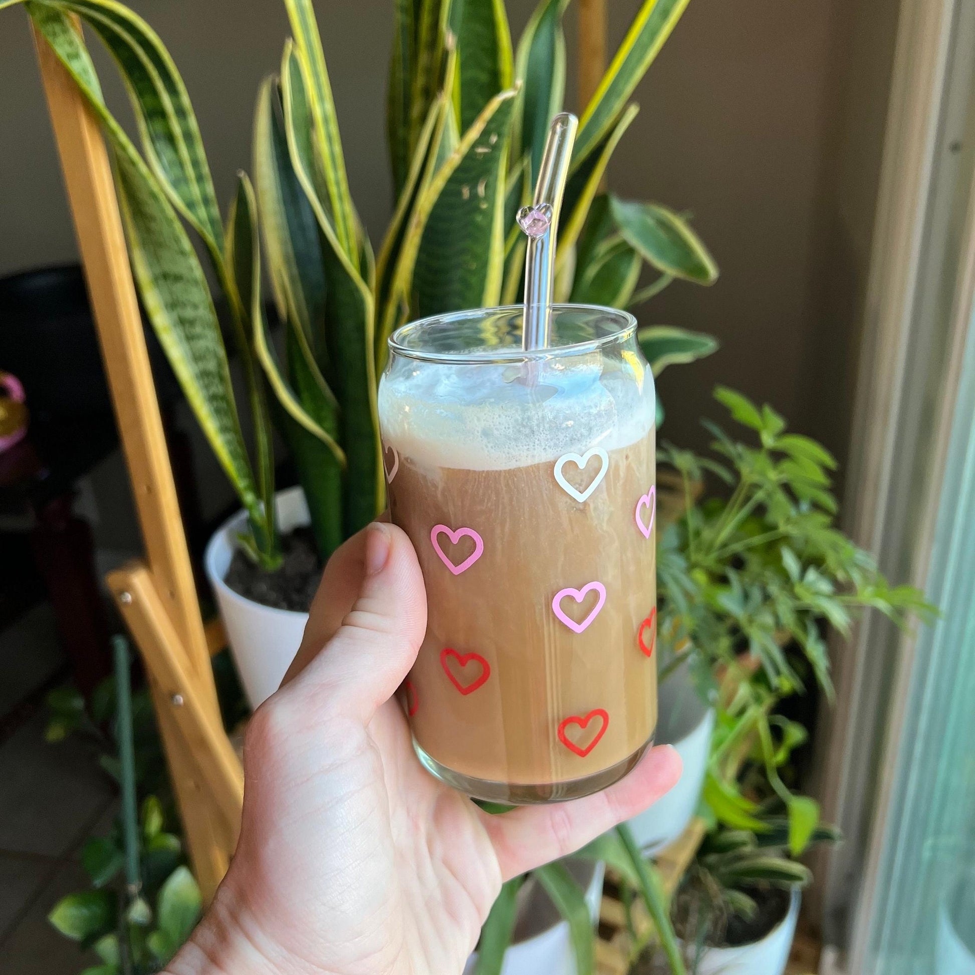 Hand holding a glass of iced coffee with white, pink and red heart designs, surrounded by plants.
