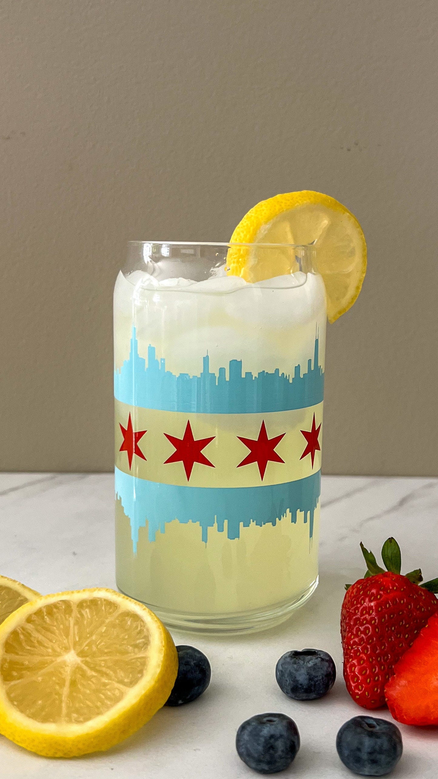 Glass with the Chicago flag with a blue skyline and red star design, lemon slice, ice cubes, and fruit on a marble surface.