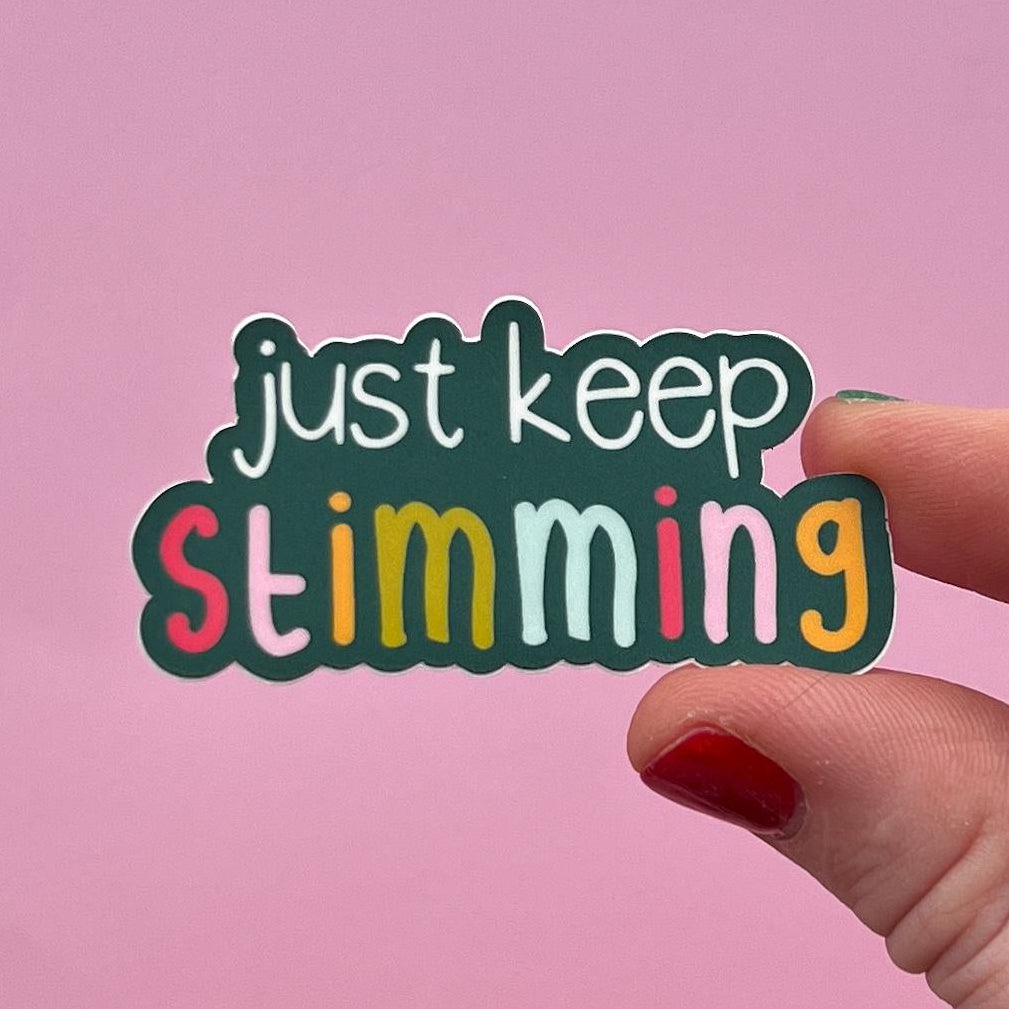 Hand holding a colorful sticker with 'just keep stimming' text against a pink background