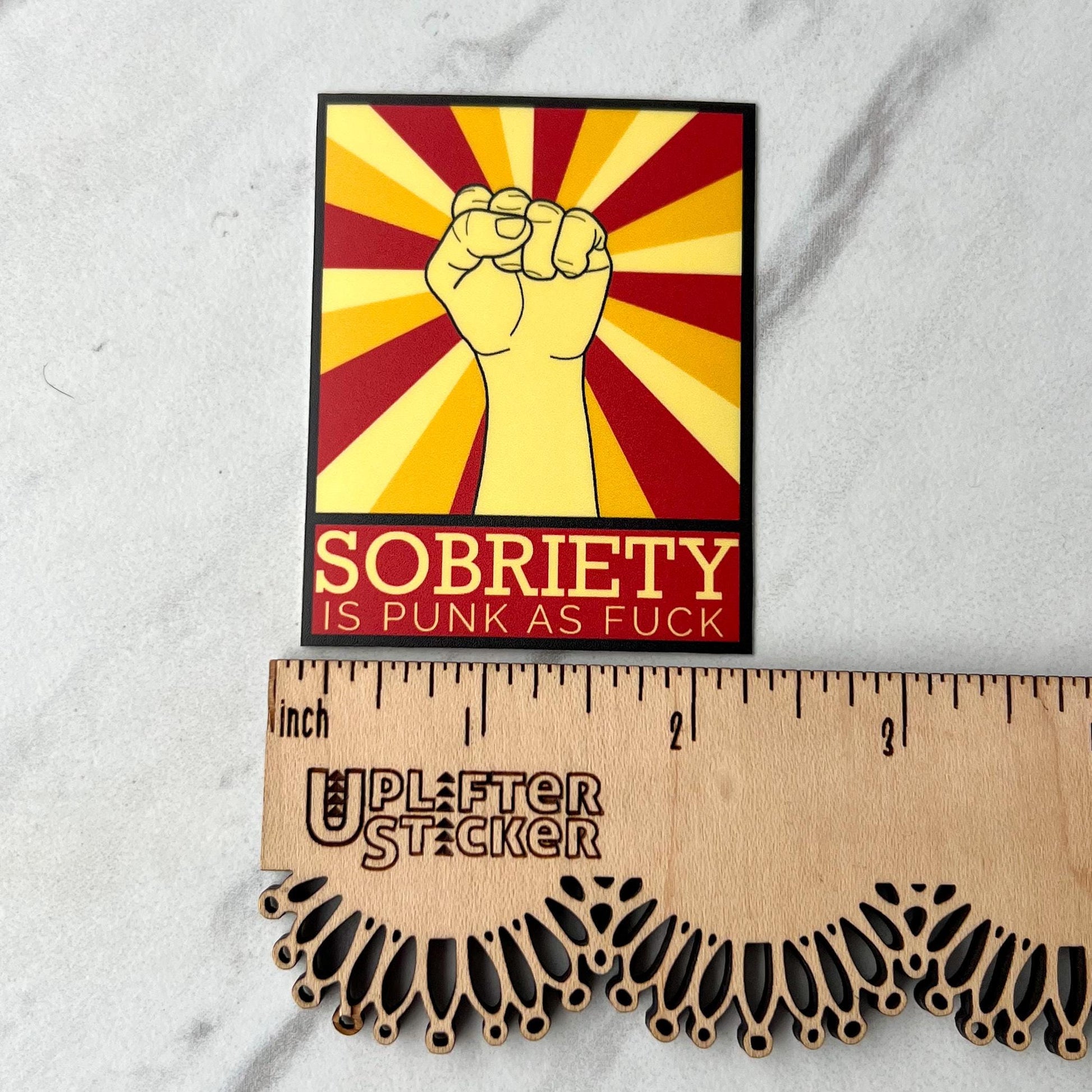 Sticker with a fist graphic and text 'Sobriety is Punk as Fuck' on a red and yellow background, next to a ruler for scale.
