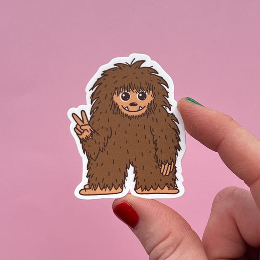 Sticker of a fluffy Bigfoot making a peace sign held by a hand against a pink background