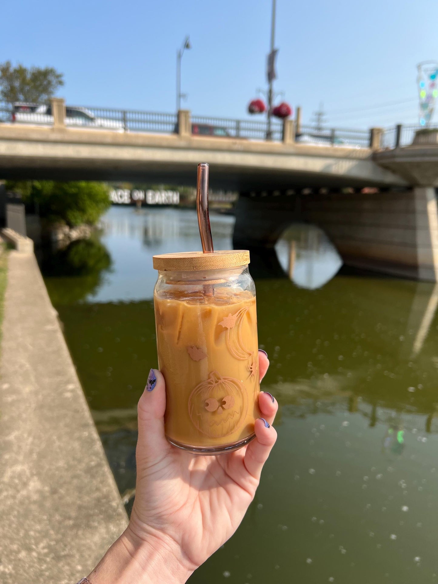 Hand holding a glass of iced coffee with a straw and halloween themed design, standing by a river with a bridge in the background.