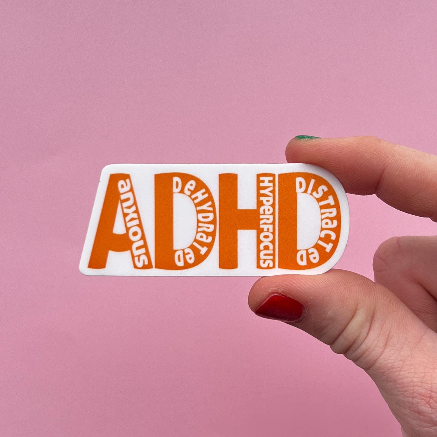 Hand holding an orange sticker that says ADHD with anxious, dehydrated, hyper focus and distracted on the letters on a pink background