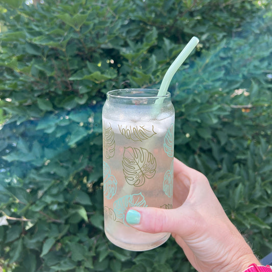 Hand holding a clear cup of water with a straw, featuring monstera pattern, against a green outdoor background.