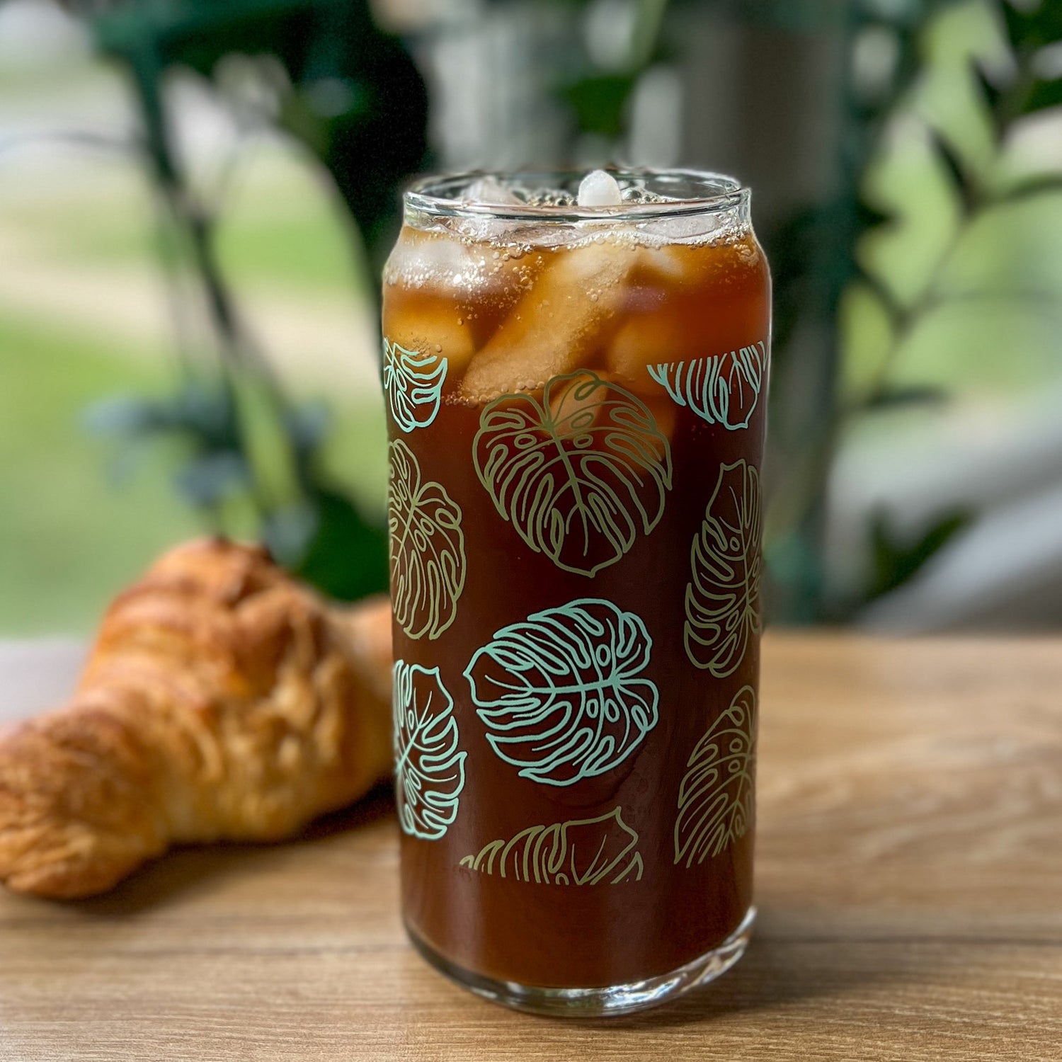 Glass of iced coffee with monstera design on a wooden table with a croissant, outdoor setting