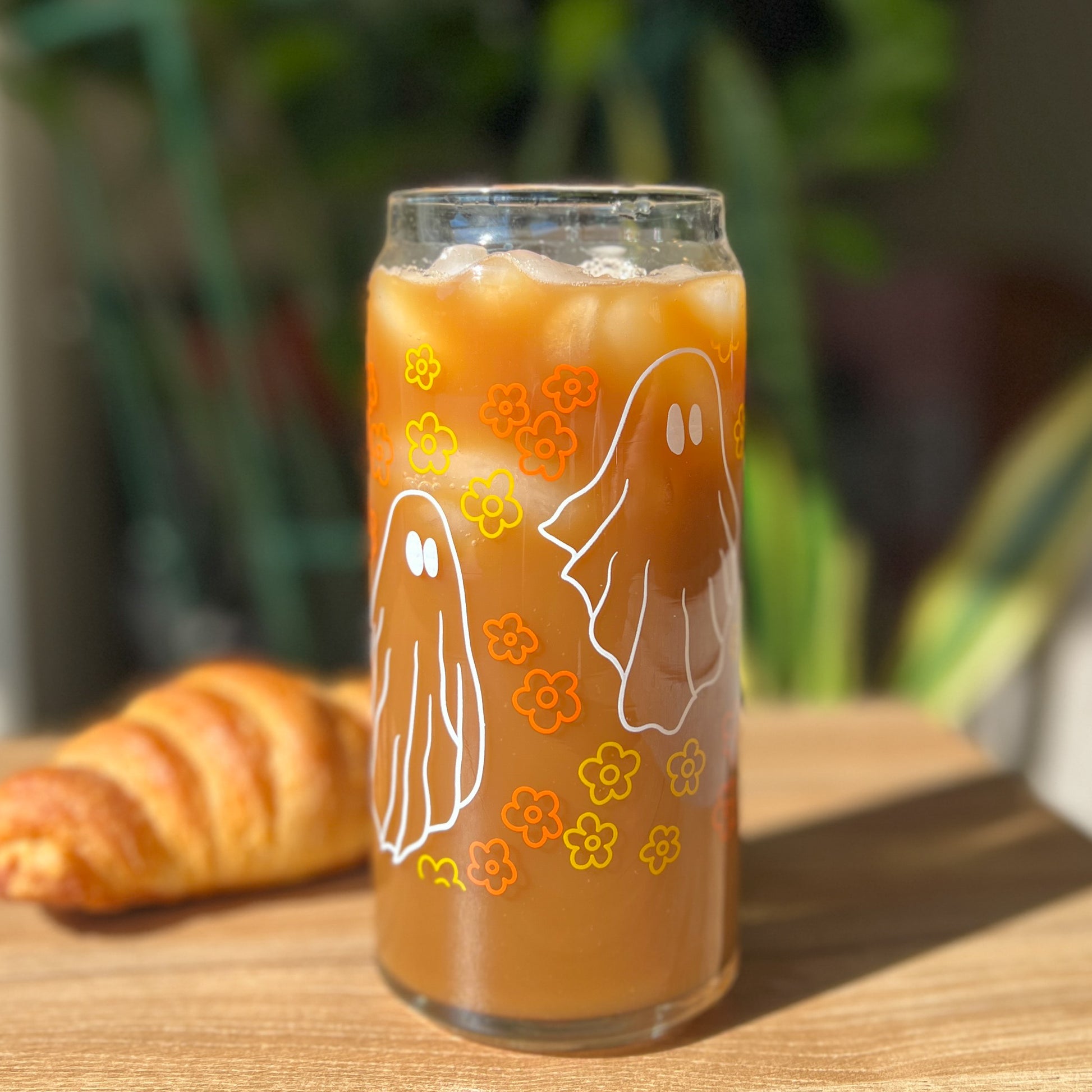 Glass of iced coffee with ghost and flower designs on a wooden table with a croissant.