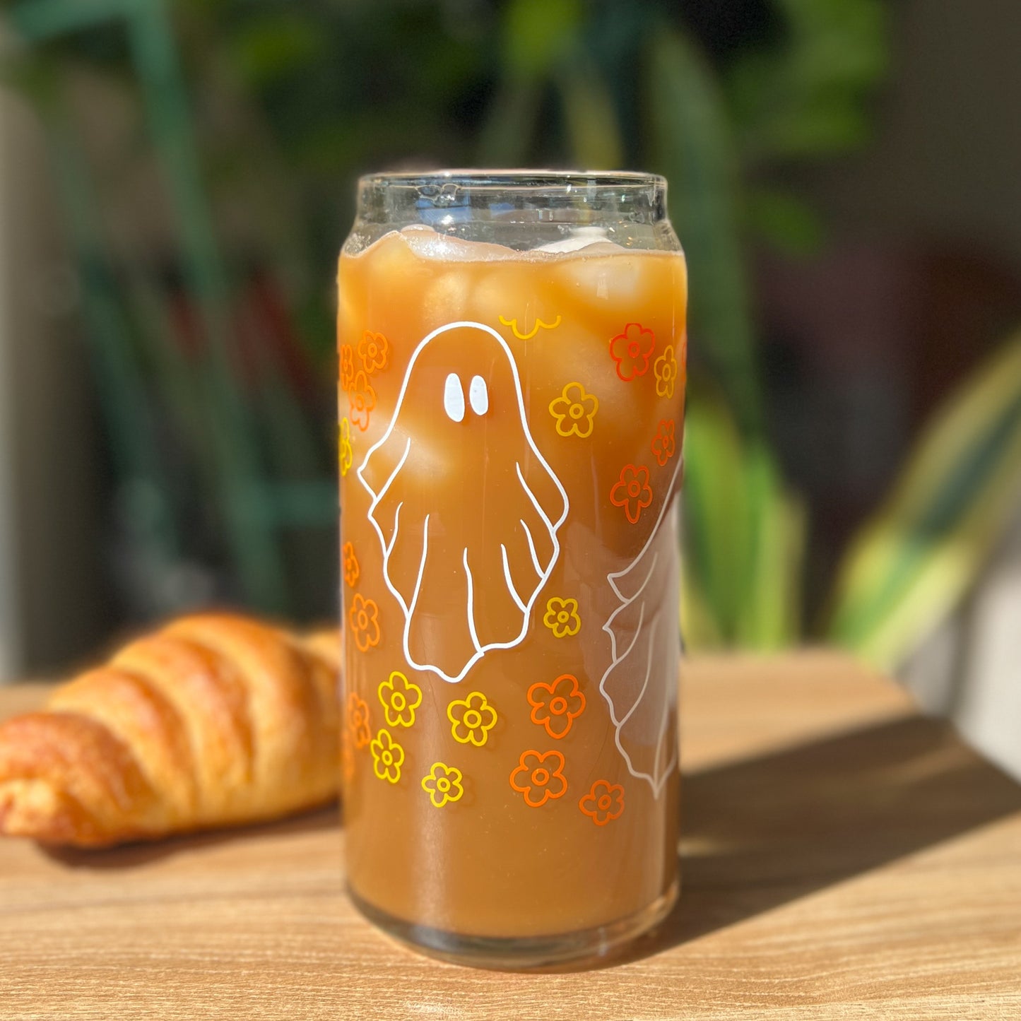 Glass of iced coffee with a ghost and vintage floral design on a wooden table with a croissant.