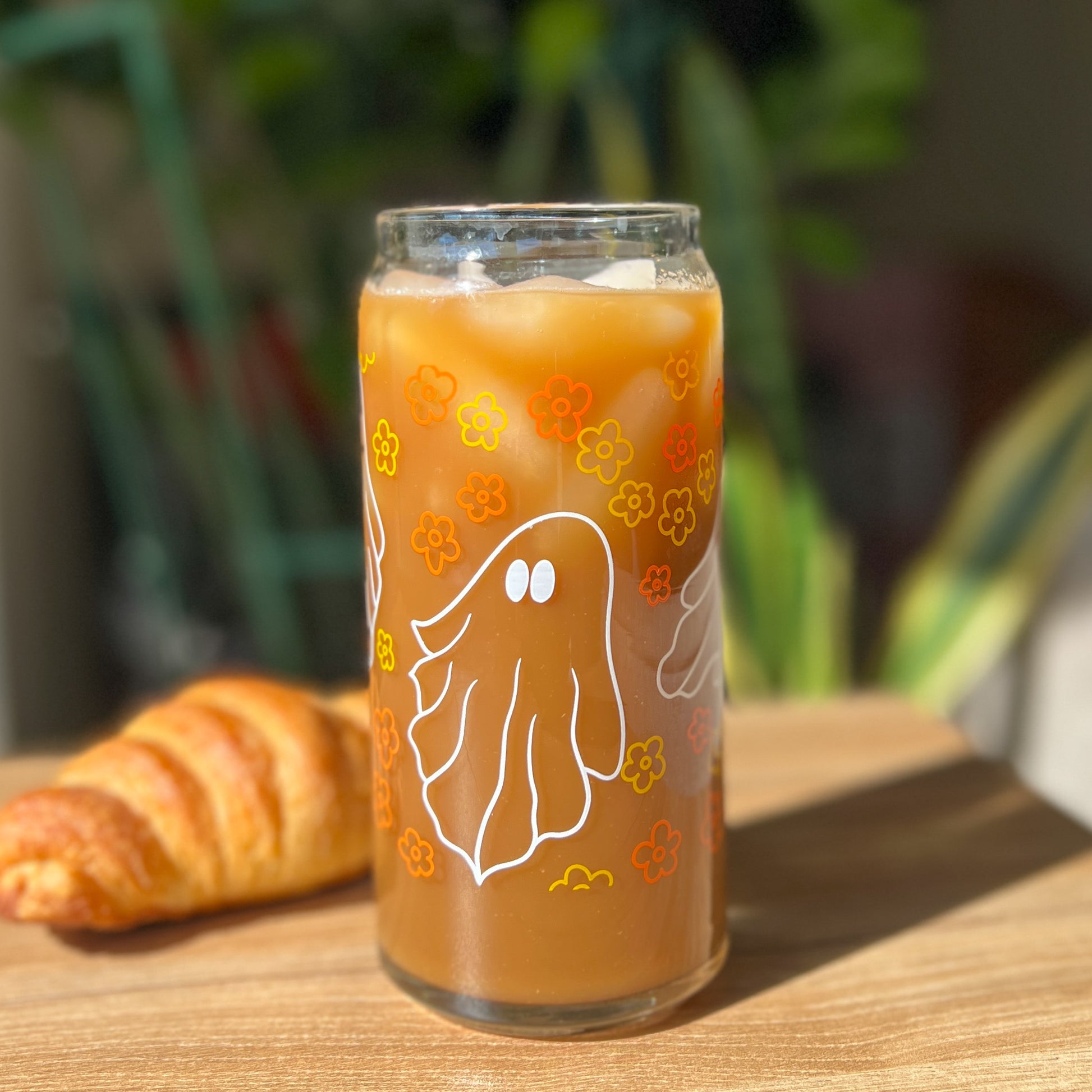 Glass of iced coffee with a ghost and vintage floral design on a wooden table with a croissant.