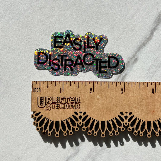 Glittery 'Easily Distracted' sticker on a gray background with a ruler for scale.