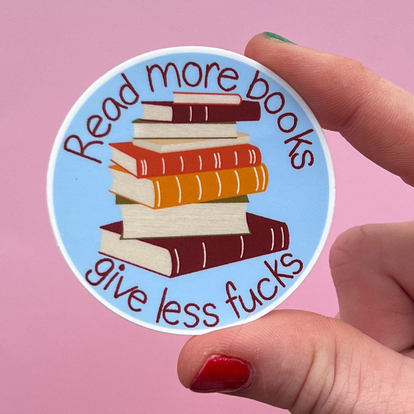 Sticker with books and text that reads 'read more books give less fucks' held by a hand against a pink background