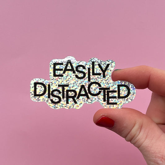 Hand holding a glittery 'EASILY DISTRACTED' sticker against a pink background