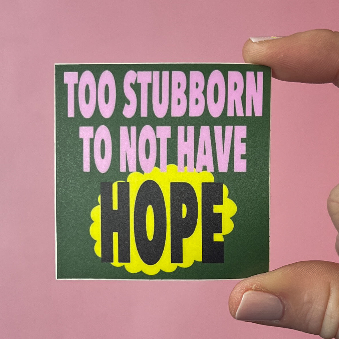 Hand holding a green sticker with motivational text 'Too stubborn to not have hope' on a pink background