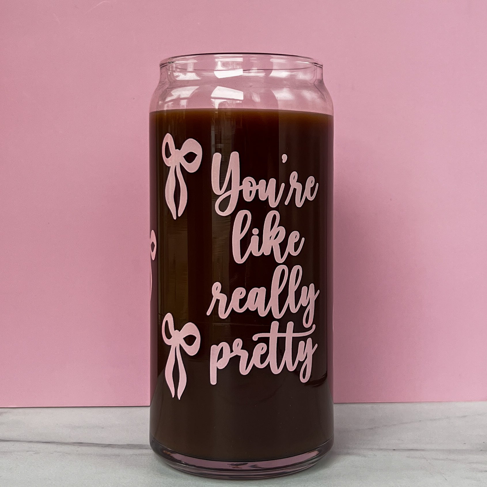 Glass cup with 'You're like really pretty' text and pink ribbons on a pink background