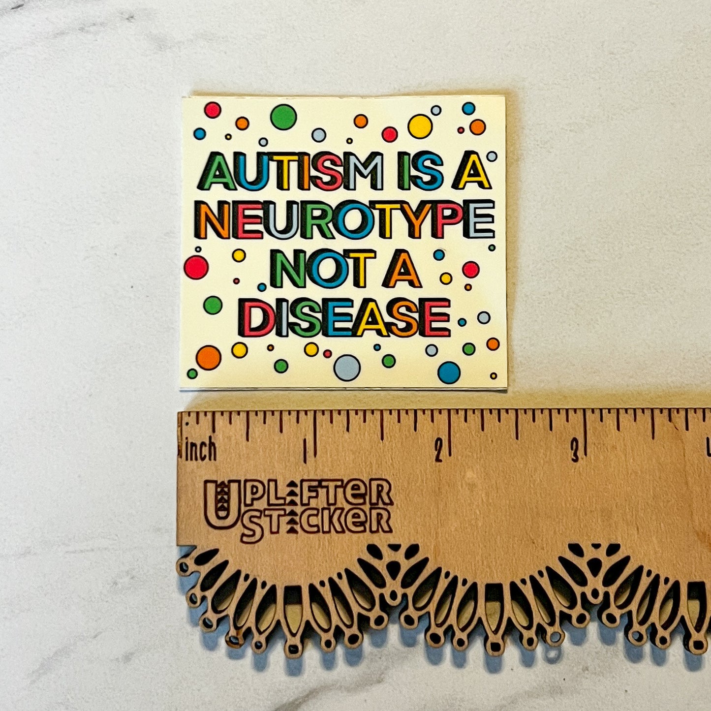 Colorful sign with 'Autism is a neurotype not a disease' text next to a wooden ruler with Uplifter Sticker branding.