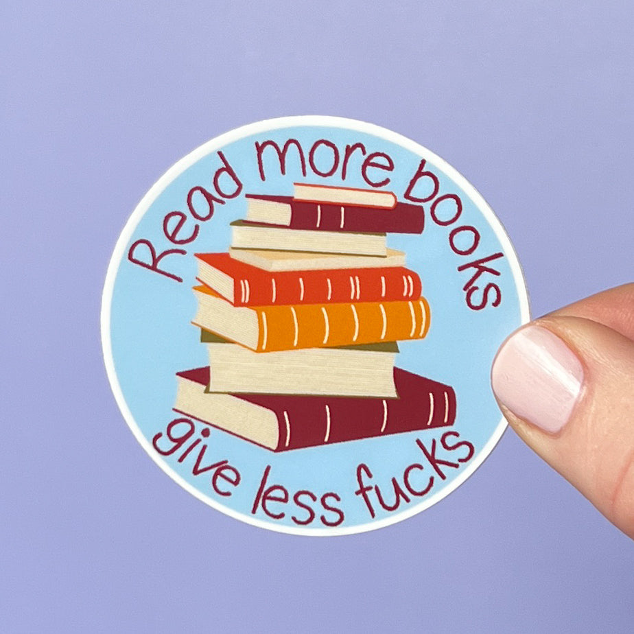 Sticker with a stack of books and text 'Read more books, give less fucks' held against a purple background.