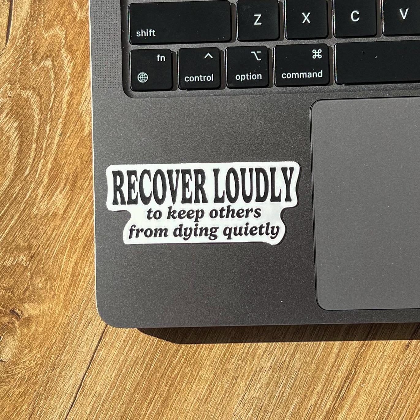 Laptop with a sticker with text 'RECOVER LOUDLY to keep others from dying quietly' on it.