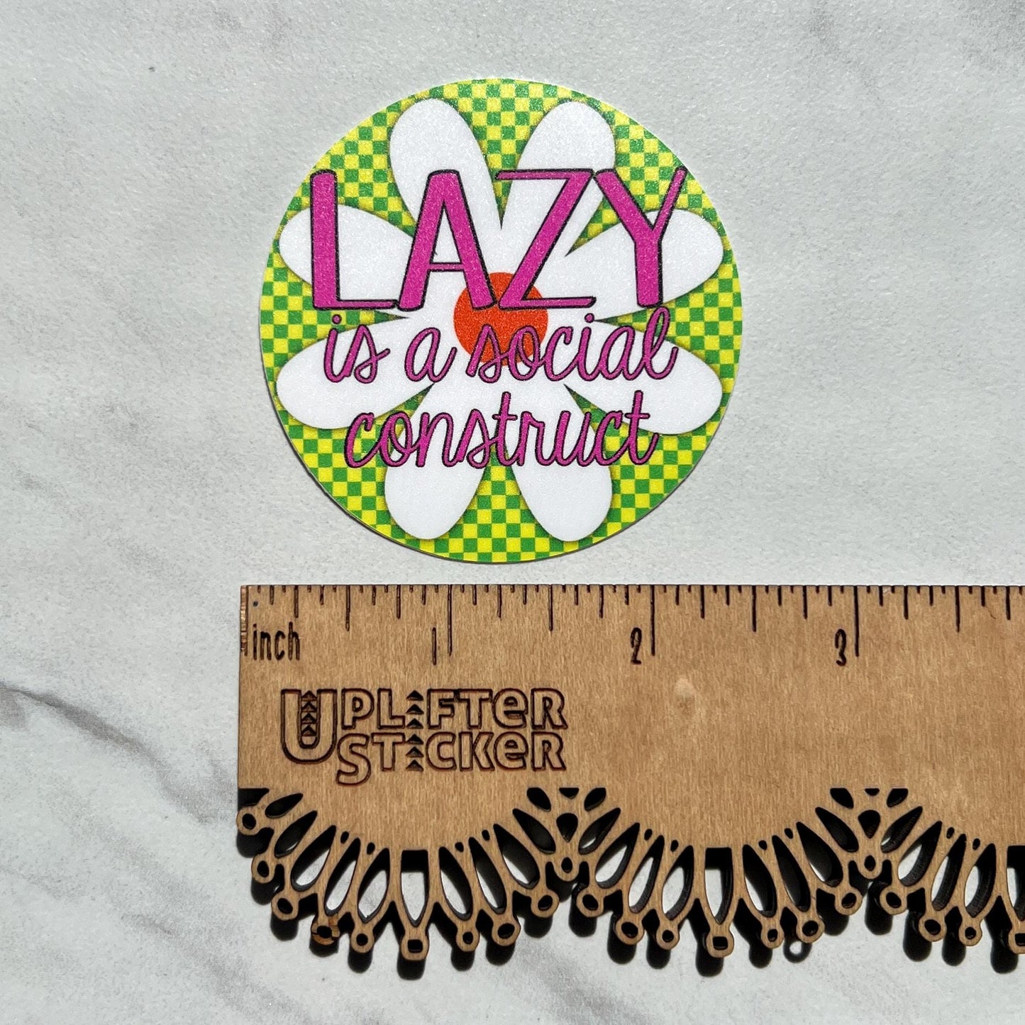 Sticker with 'LAZY is a social construct' text on a green checkered background, ruler for scale, and Uplifter Sticker branding.
