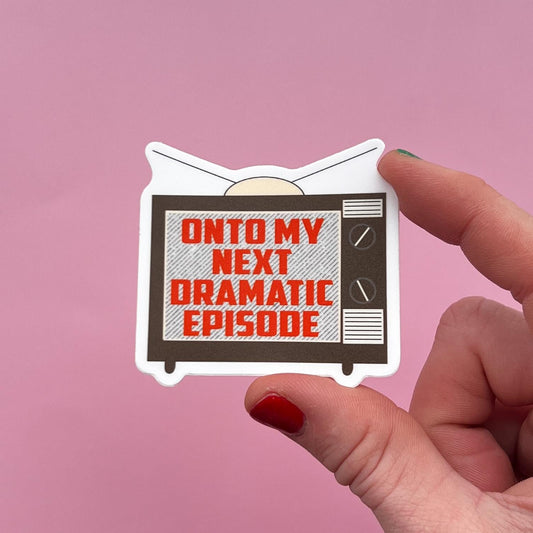 Hand holding a sticker with a retro TV sticker and text 'Onto my next dramatic episode' on a pink background.