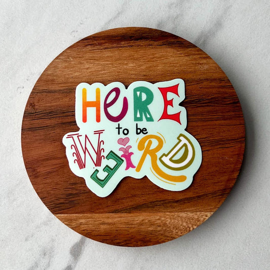 Round wooden coaster with colorful sticker and text that says 'here to be weird' on a light gray background
