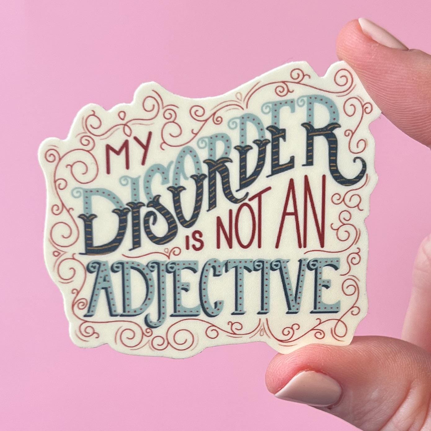 Hand holding a decorative sticker with text  that reads 'my disorder is not an adjective' on a pink background
