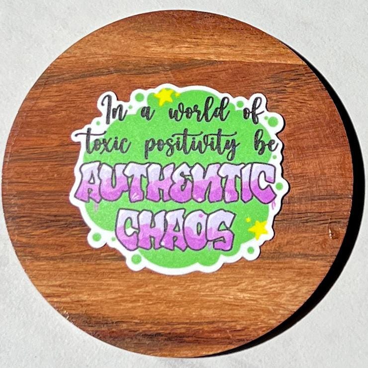 Sticker with 'In a world of toxic positivity be Authentic Chaos' text on a wooden surface