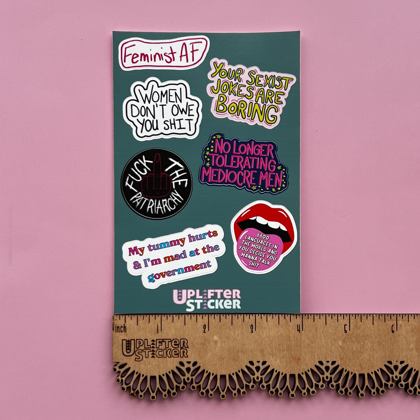 Collection of politically charged stickers on a teal card with a ruler for scale, against a pink background.