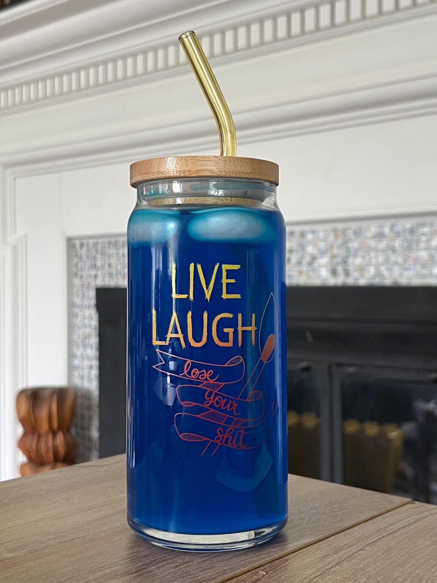 Blue glass jar with text that reads 'live laugh lose your shit' with wooden lid and gold straw, containing a blue liquid, on a wooden surface.