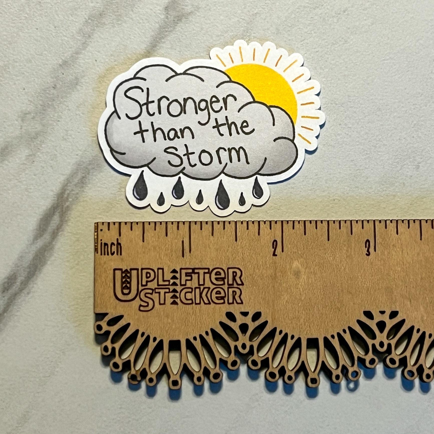 Sticker with 'Stronger than the Storm' text on a cloud with raindrops and sun, placed on a wooden ruler.