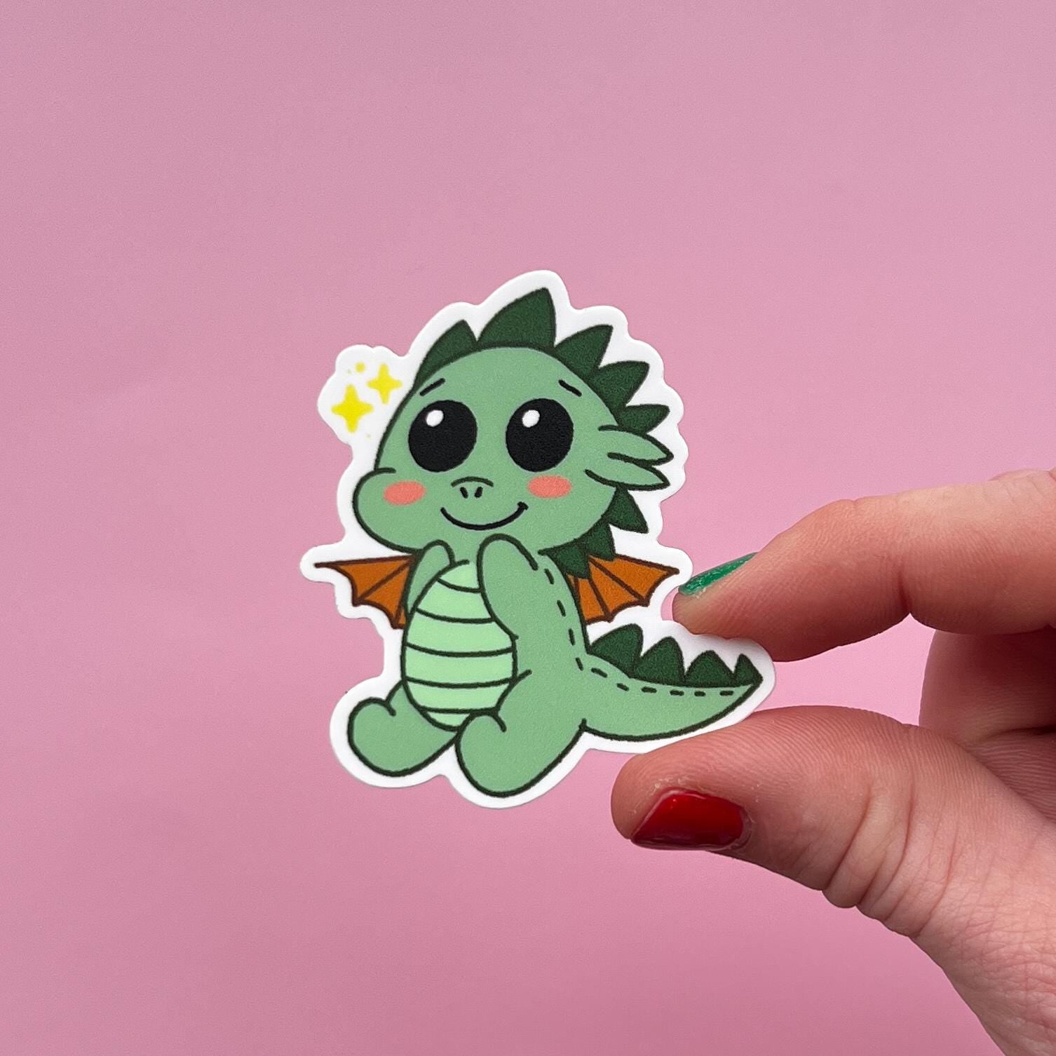 Hand holding a cute green dragon sticker against a pink background