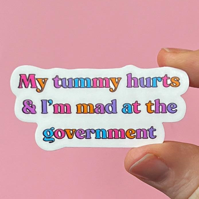 Sticker with colorful text 'My tummy hurts & I'm mad at the government' held against a pink background