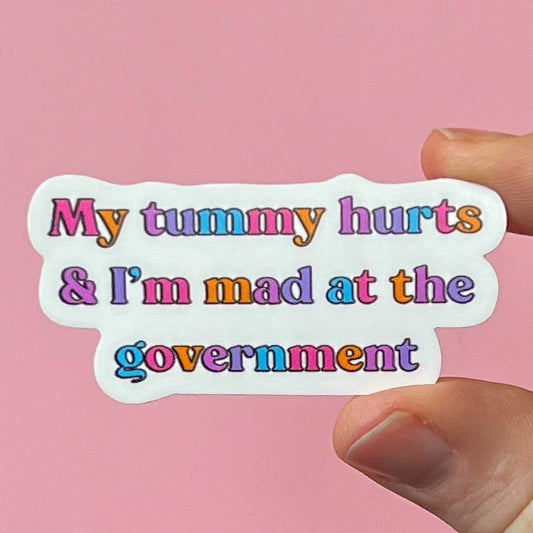 Sticker with colorful text 'My tummy hurts & I'm mad at the government' held against a pink background