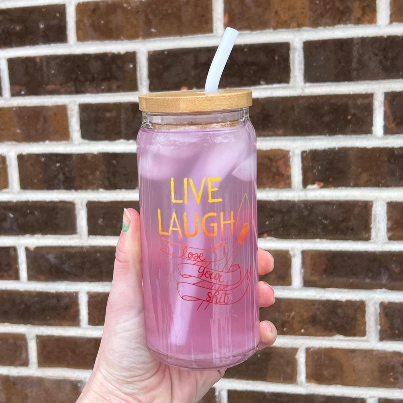 Hand holding a pink glass tumbler with 'Live Laugh Love your self' text against a brick wall.