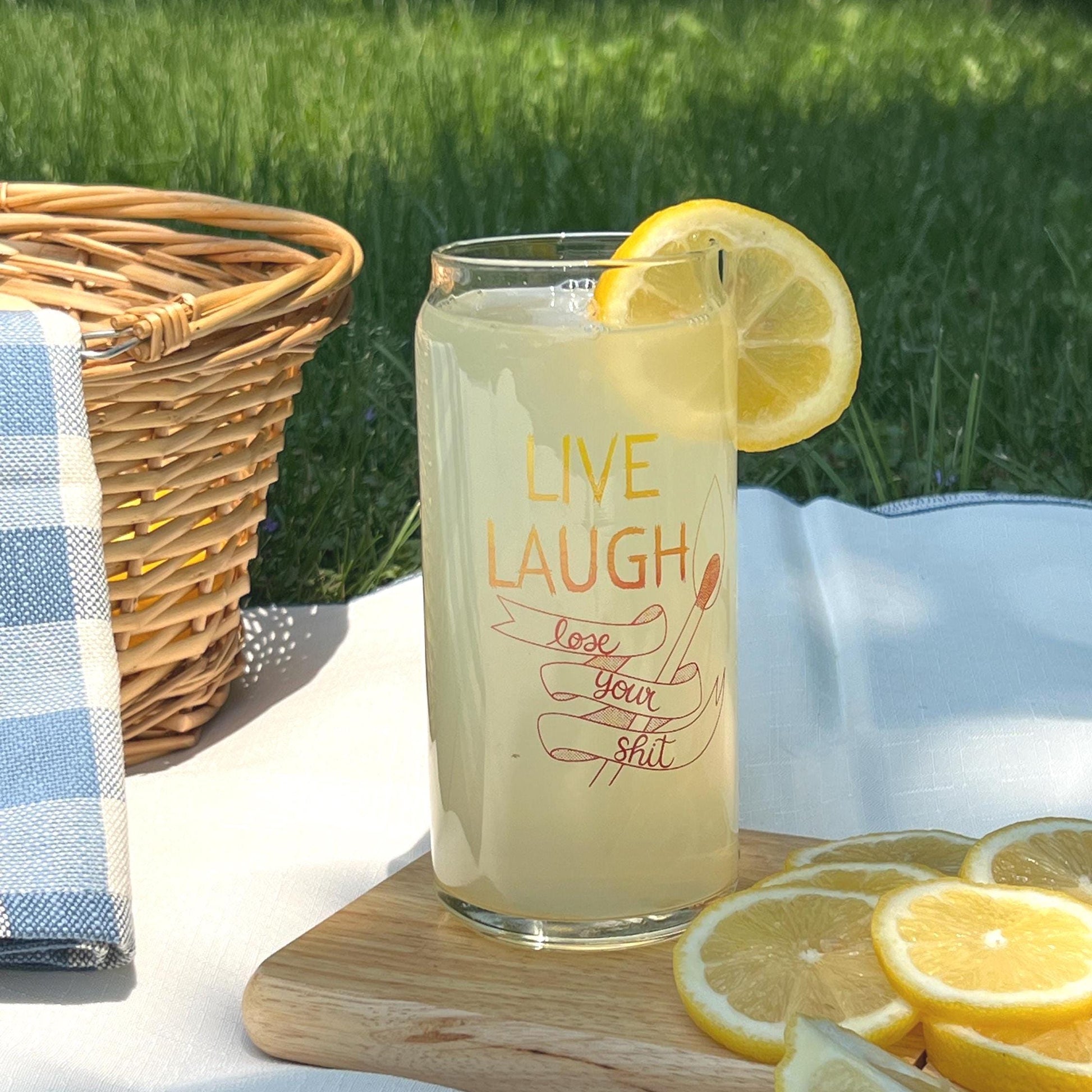 Glass of lemonade with a lemon slice, text 'Live Laugh Lose Your Shit', on a picnic setting.