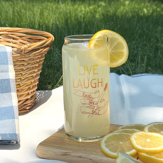 Glass of lemonade with a lemon slice, text 'Live Laugh Lose Your Shit', on a picnic setting.