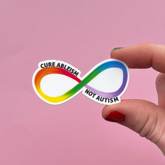 Sticker with rainbow infinity symbol and text 'Cure Ableism Not Autism' held by a hand on a pink background