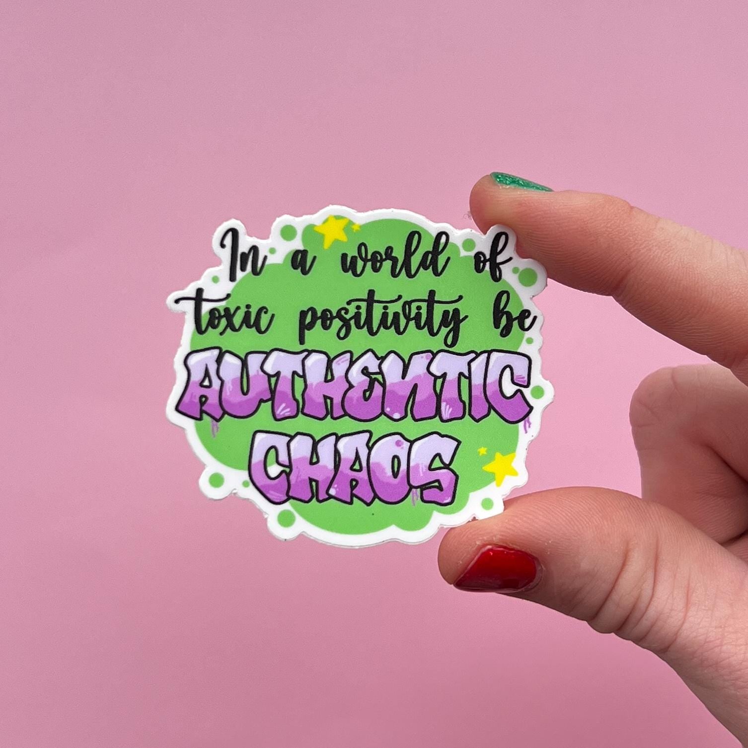 Hand holding a colorful sticker stating "in a world of toxic positivity be authentic chaos" against a pink background