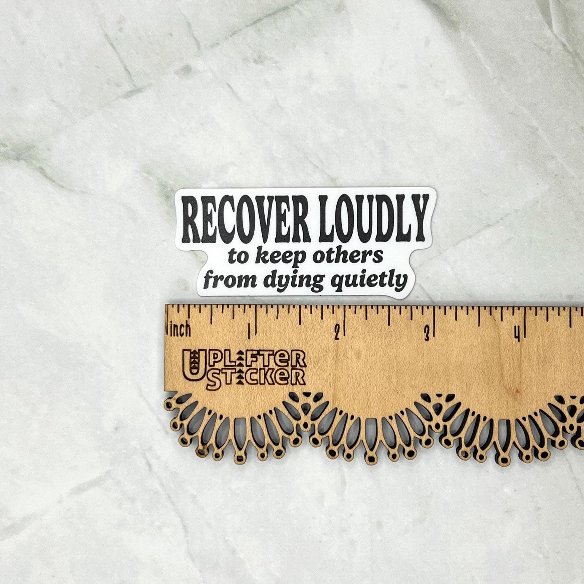 Sticker with text 'RECOVER LOUDLY to keep others from dying quietly' on a marble surface with a ruler for scale.
