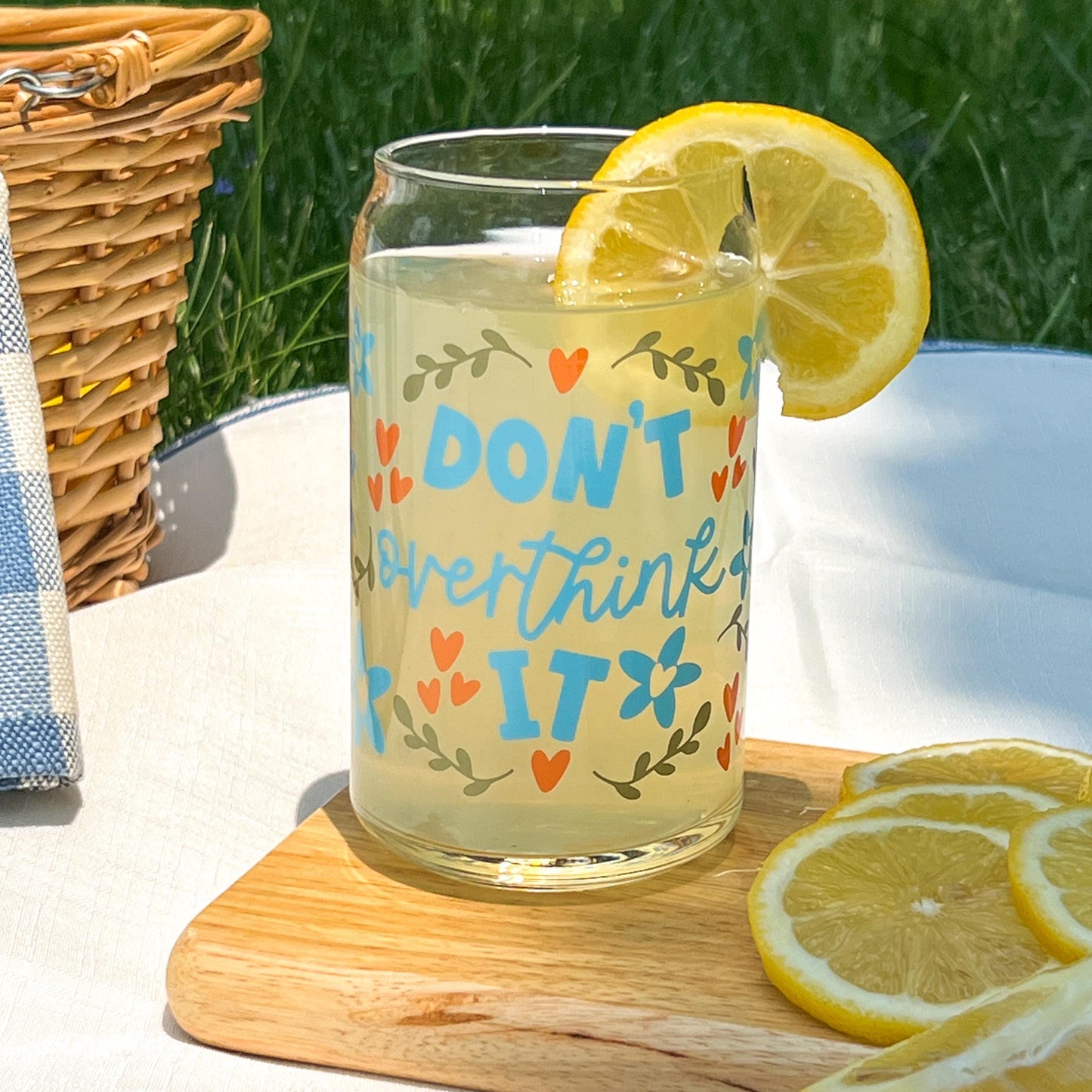Glass of lemonade with a lemon slice on a wooden coaster outdoors. Cup says "don't overthink it"