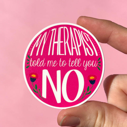A round sticker with a pink background featuring the text 'MY THERAPIST TOLD ME TO TELL YOU NO' along with some decorative elements like flowers and leaves. The sticker is held in a hand against a pink background.