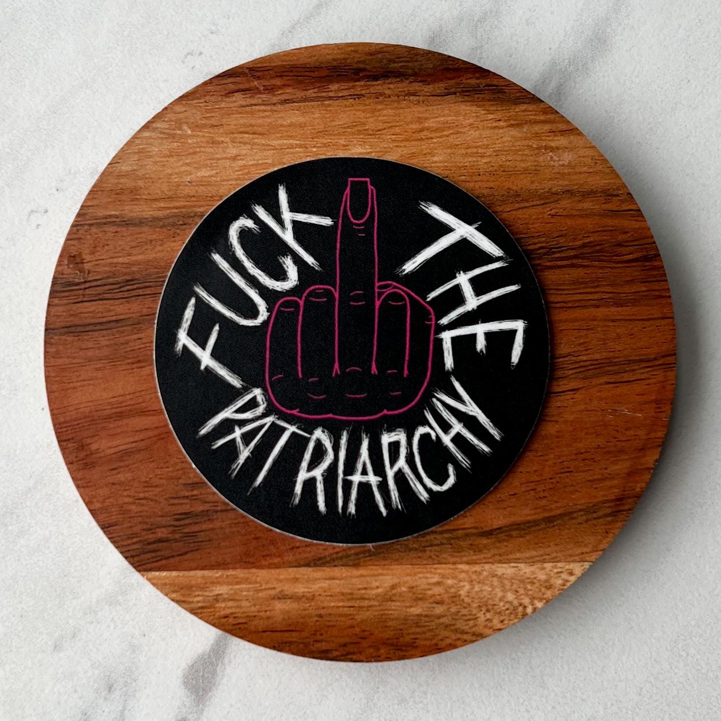 Round wooden sign with a middle finger graphic and fuck the patriarchy  text on a white background
