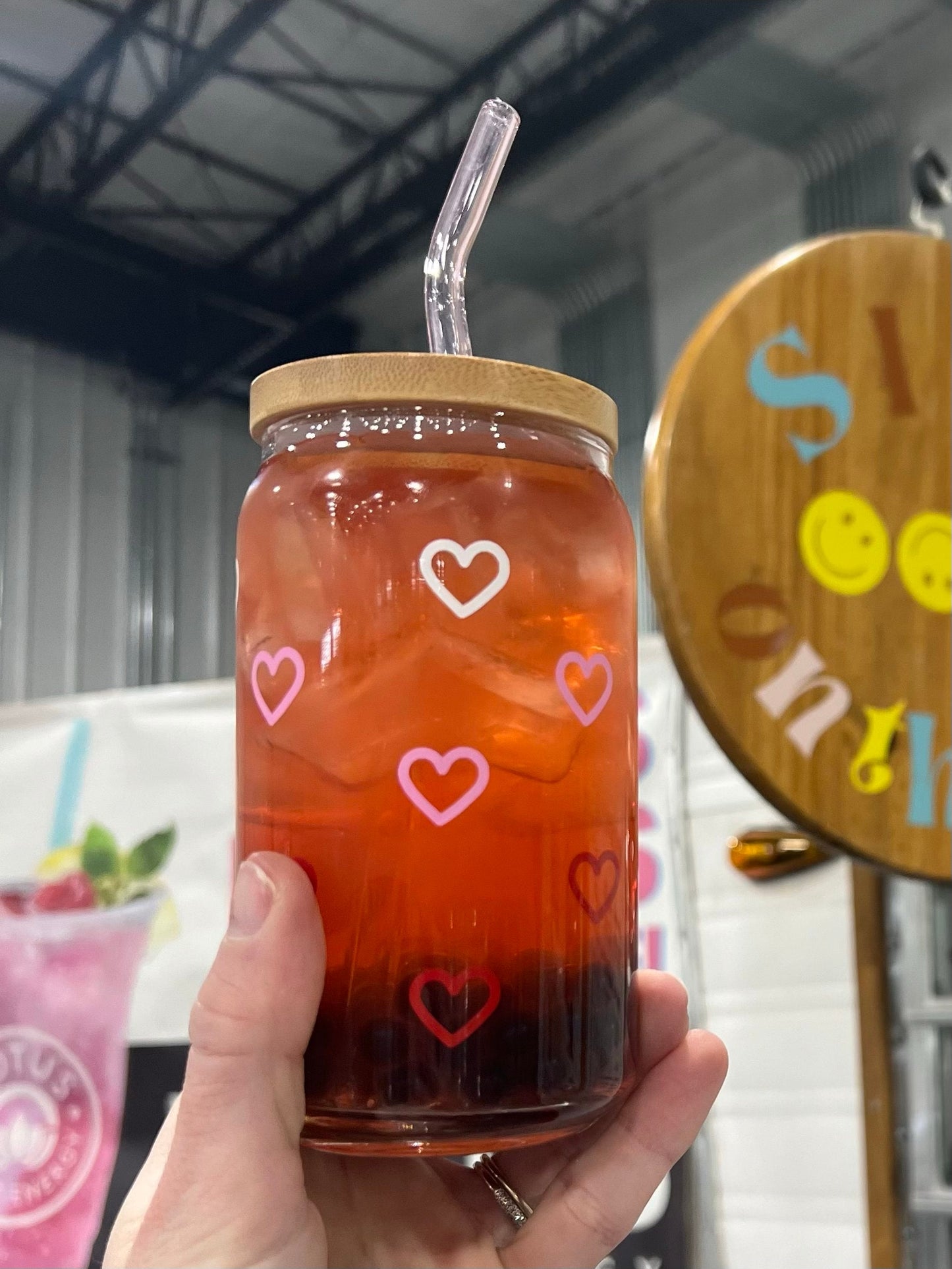 Hand holding a jar of iced tea with white, pink, and red heart designs against a blurred indoor background