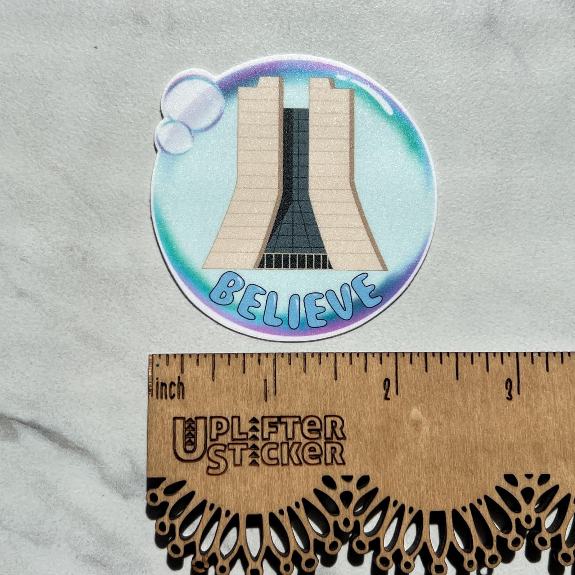 Sticker with a design of Fermilab and the word 'BELIEVE' in a bubble on a gray background, next to a ruler for scale.