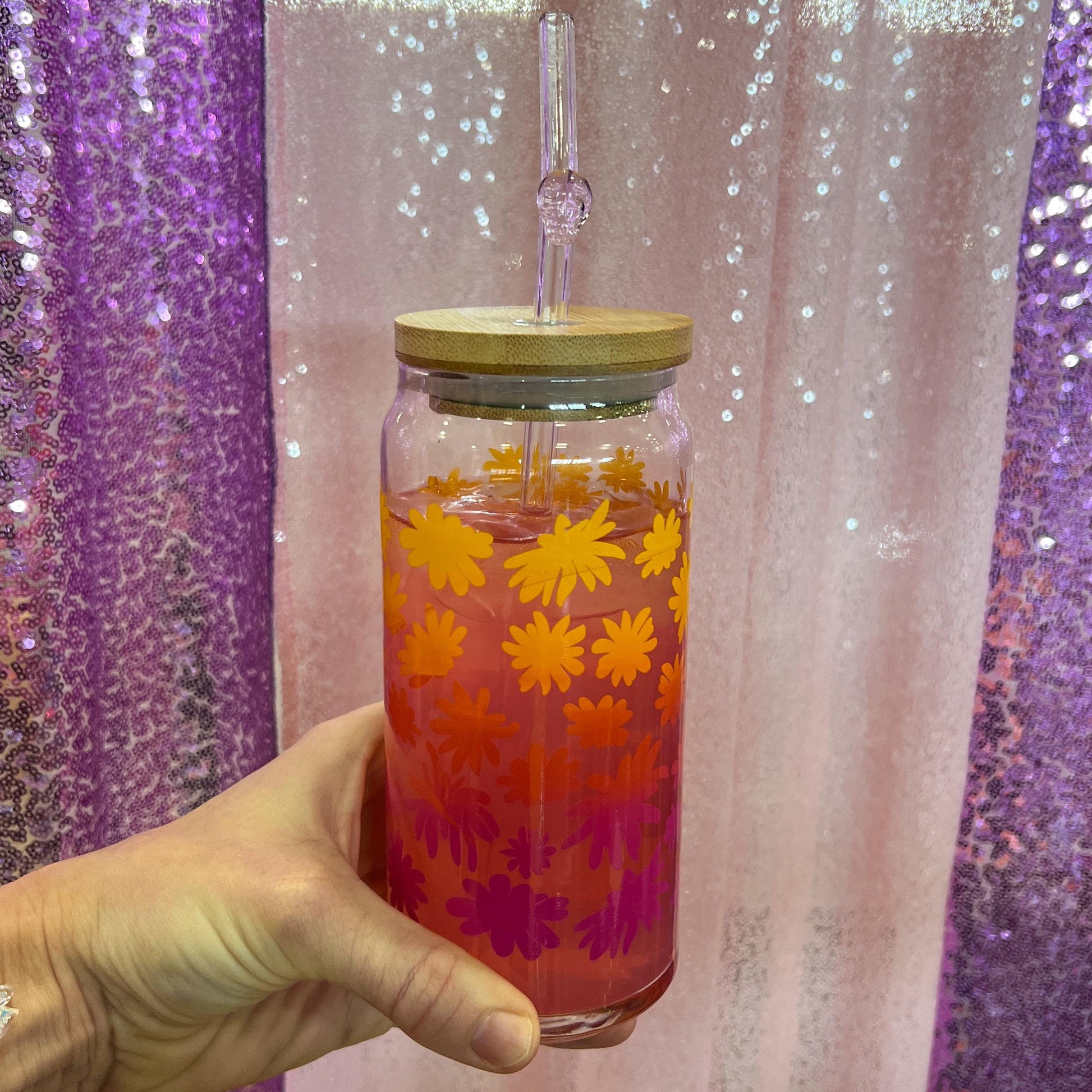 Hand holding a glass jar with floral pattern against a glittery purple and pink background