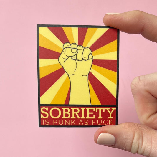 Sticker with a fist and text 'Sobriety is punk as fuck' held by a hand against a pink background